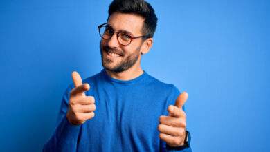 Jeune homme beau avec barbe portant un pull et des lunettes sur fond bleu pointant des doigts vers la caméra