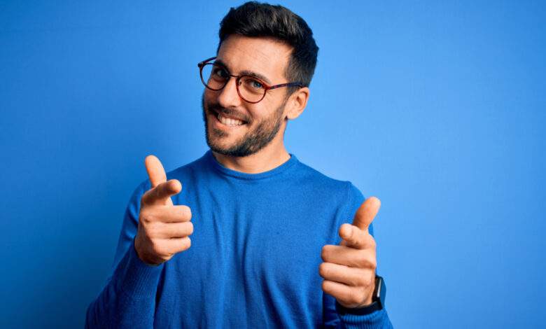 Jeune homme beau avec barbe portant un pull et des lunettes sur fond bleu pointant des doigts vers la caméra
