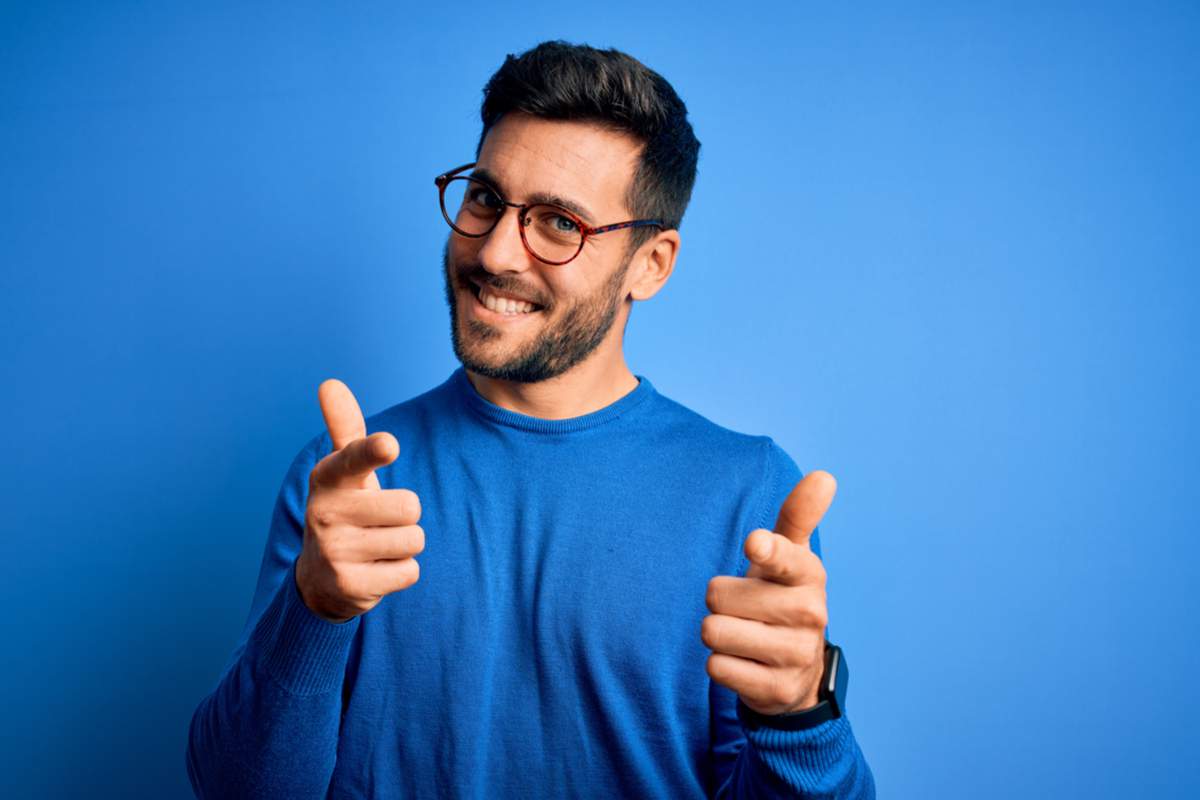 Jeune homme beau avec barbe portant un pull et des lunettes sur fond bleu pointant des doigts vers la caméra