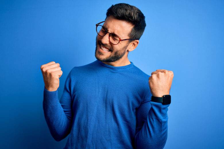 Jeune homme beau avec barbe portant un pull et des lunettes sur fond bleu très heureux et excité