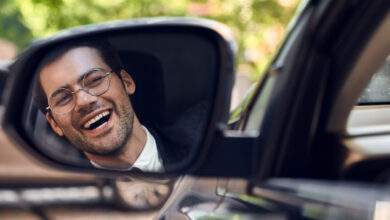 Un homme avec un large sourire au visage regarde dans le rétroviseur latéral de sa voiture.