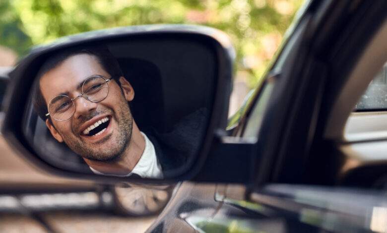 Un homme avec un large sourire au visage regarde dans le rétroviseur latéral de sa voiture.