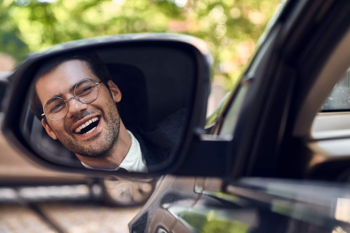 Un homme avec un large sourire au visage regarde dans le rétroviseur latéral de sa voiture.