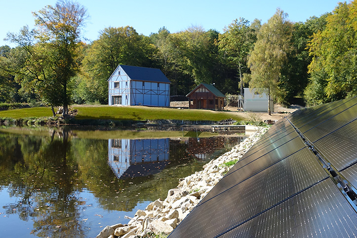 Une maison passive bleue au bord d'un lac avec des panneaux solaires.