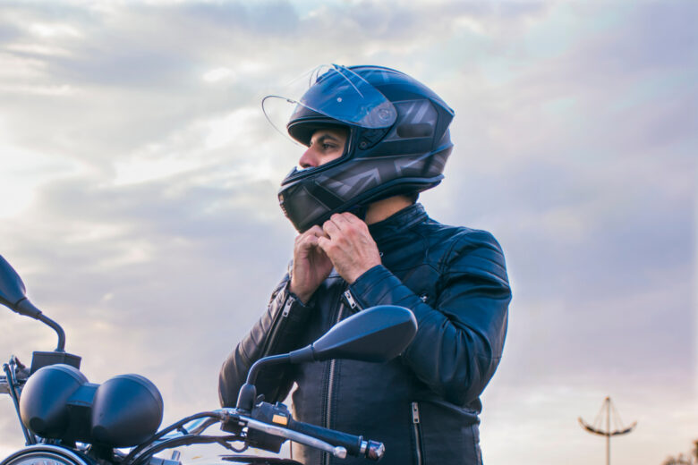 Homme assis sur une moto, portant un jean et une veste noire, attachant son casque