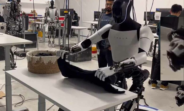 Le robot Optimus de Tesla pliant du linge, on distingue sur la droite la main d'un opérateur contrôlant cette tâche.