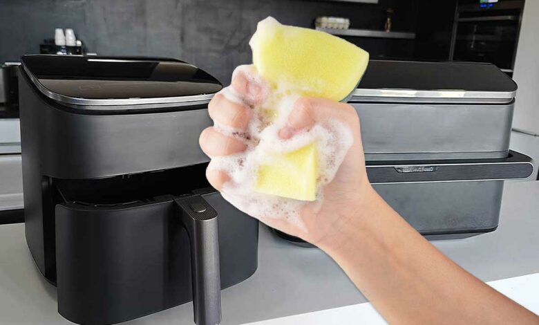Une main tenant une éponge devant deux airfryers.